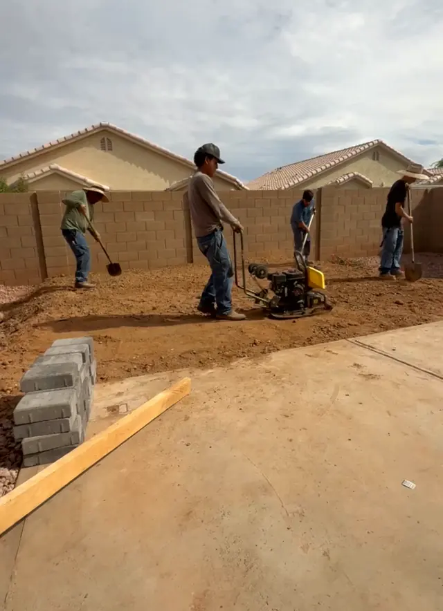 Gio and his crew compacting the ground of a backyard with a plate compactor, shovels in hand, in front of a block wall